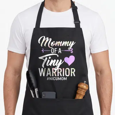 Mother tiny chef apron - Stacked floral text Mommy Of A Tiny Warrior with purple arrow heart and NICU hashtag — shown on a black cotton canvas apron against a white background