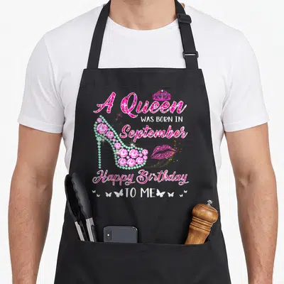 Funny september baking queen apron - Jeweled pink high heel with A Queen Was Born In September text and butterflies — shown on a black cotton canvas apron against a white background