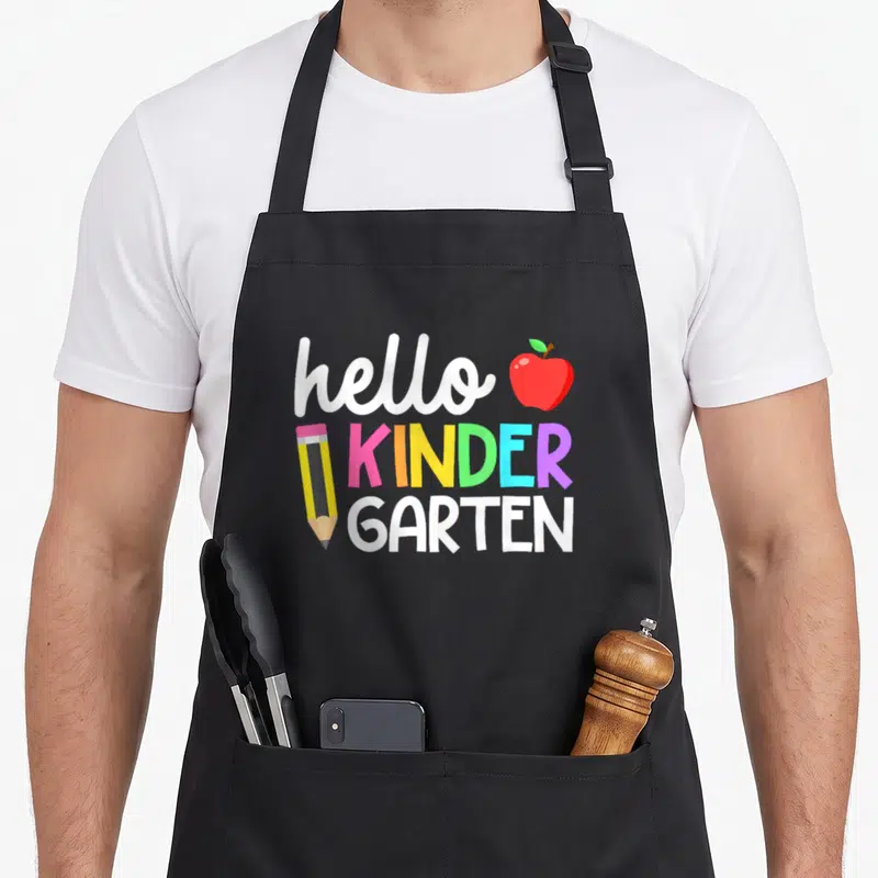 Hello cute teacher aprons - Hello Kinder Garten text with red apple and yellow pencil in cartoon style — shown on a black cotton canvas apron against a white background