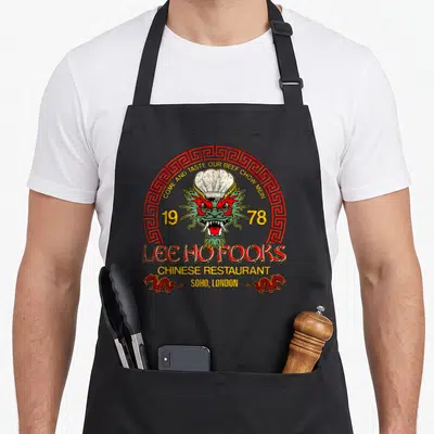 Fook chinese apron - Retro Lee Ho Fooks dragon chef hat 1978 Soho London text red yellow green — shown on a black cotton canvas apron against a white background
