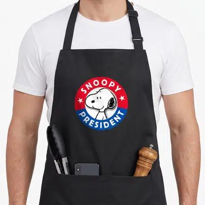 Funny presidents apron - Snoopy face inside red white and blue circular campaign emblem with text — shown on a black cotton canvas apron against a white background