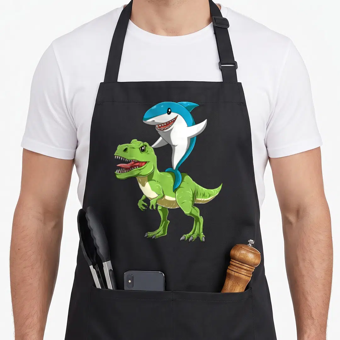 Jawsome dinosaur apron - Blue shark riding on back of green tyrannosaurus rex cartoon — shown on a black cotton canvas apron against a white background
