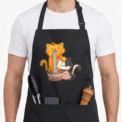Ramen anime kawaii aprons - Orange cat eating noodles from bowl next to black and white cat with fish cake on head — shown on a black cotton canvas apron against a white background