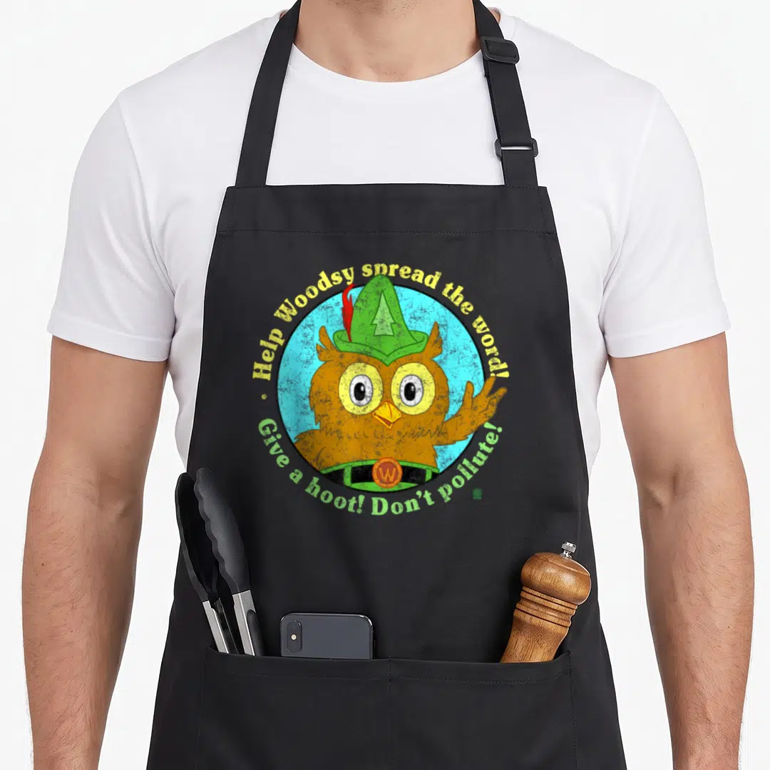 Pollute owl apron - Cartoon brown owl in green hat inside blue circle with yellow and green text — shown on a black cotton canvas apron against a white background