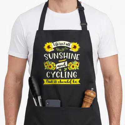 Sunshine grandma apron - Sunflowers and stacked text reading life isn't all sunshine and cycling but it should be — shown on a black cotton canvas apron against a white background
