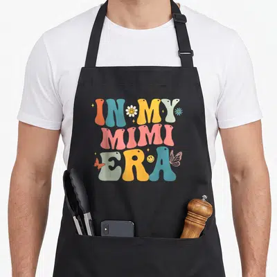 Groovy mother's day aprons - Retro wavy text IN MY MIMI ERA with colorful flowers butterflies and smiley faces — shown on a black cotton canvas apron against a white background