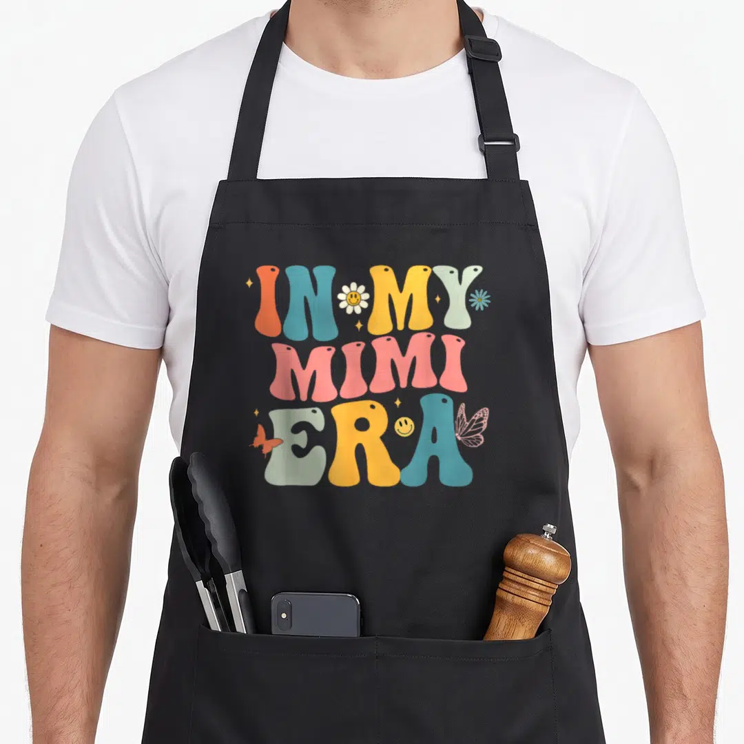 Groovy mother's day aprons - Retro wavy text IN MY MIMI ERA with colorful flowers butterflies and smiley faces — shown on a black cotton canvas apron against a white background