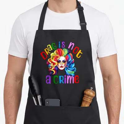Drag pride apron - Rainbow colored drag queen face with arched text Drag is not a crime below — shown on a black cotton canvas apron against a white background