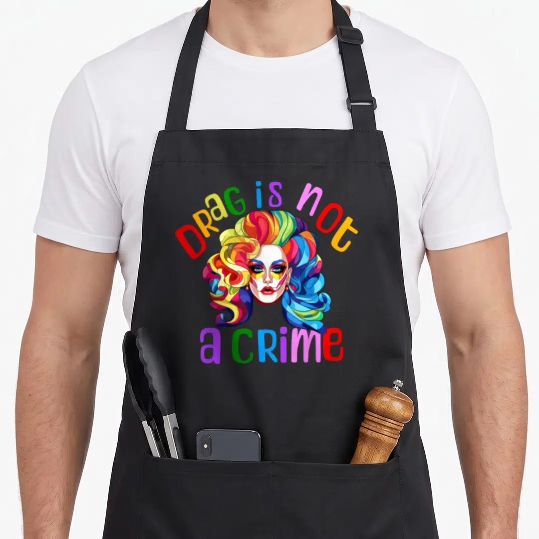 Drag pride apron - Rainbow colored drag queen face with arched text Drag is not a crime below — shown on a black cotton canvas apron against a white background