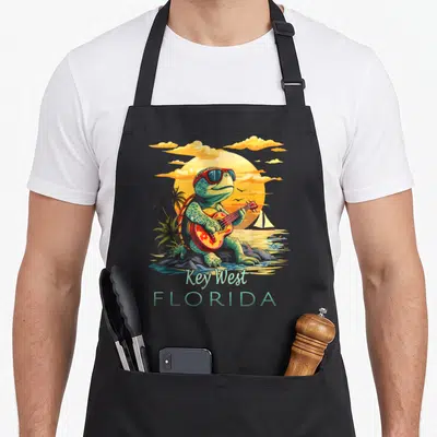Turtle florida apron - Cartoon turtle playing guitar on rocks with Key West Florida text and sunset sailboat background — shown on a black cotton canvas apron against a white background