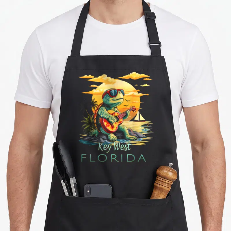 Turtle florida apron - Cartoon turtle playing guitar on rocks with Key West Florida text and sunset sailboat background — shown on a black cotton canvas apron against a white background