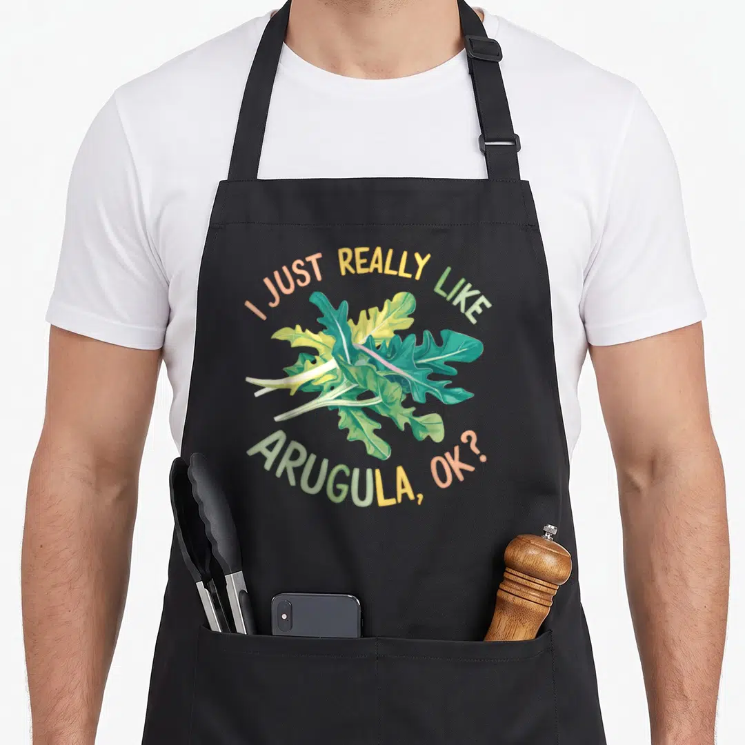 Funny arugula apron - Cartoon arugula bunch with I Just Really Like Arugula OK text in green and peach — shown on a black cotton canvas apron against a white background