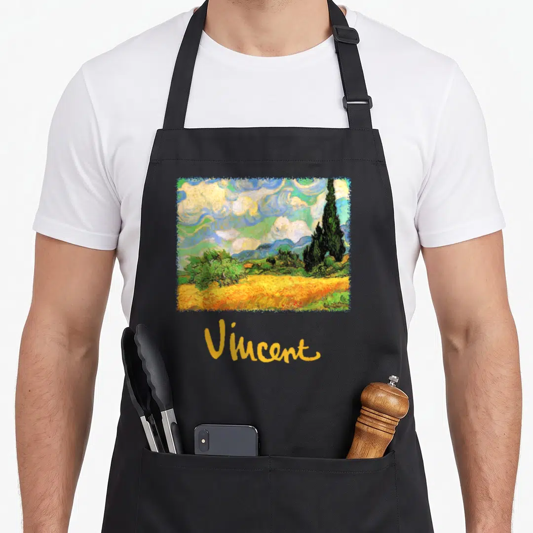 Wheat van gogh apron - Rectangular watercolor landscape with yellow fields and blue sky above the name Vincent — shown on a black cotton canvas apron against a white background