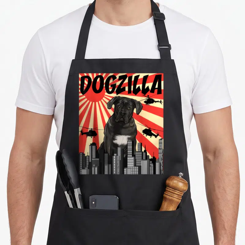 Japanese cane apron - Giant black dog and DOGZILLA text above grey skyline with helicopters on red and cream sunburst — shown on a black cotton canvas apron against a white background