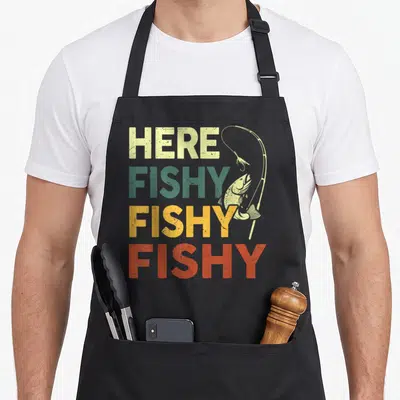 Fishy bass fishing aprons - Stacked HERE FISHY FISHY FISHY text with a hooked bass and rod — shown on a black cotton canvas apron against a white background