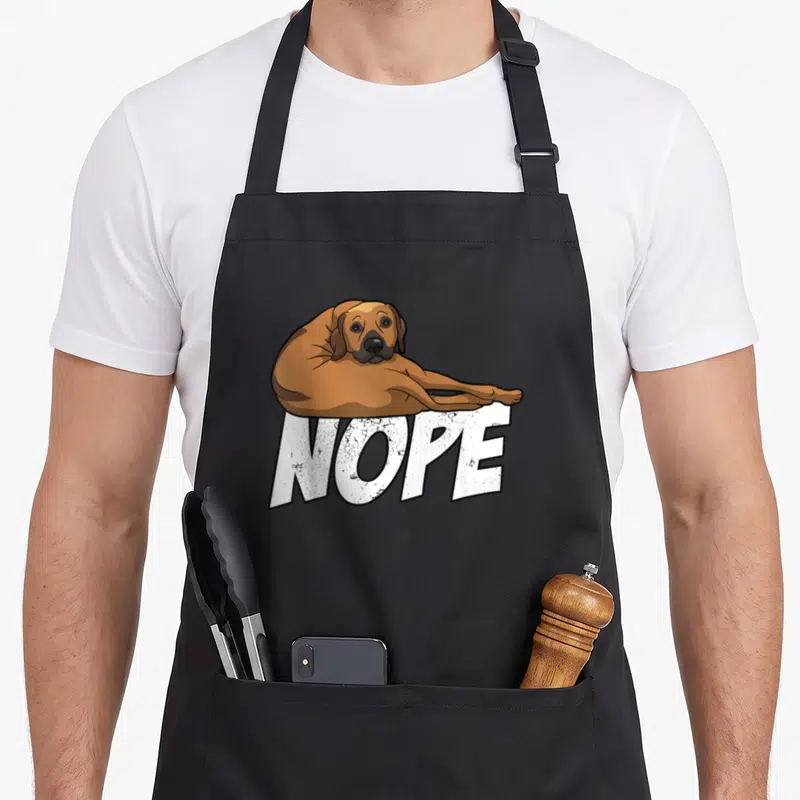 Lazy rhodesian dog apron - Cartoon brown dog lying on white NOPE text on black apron — shown on a black cotton canvas apron against a white background
