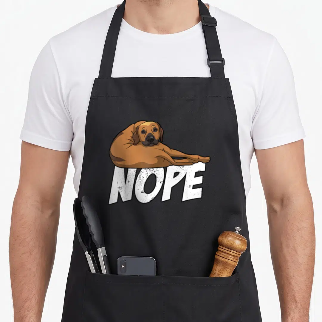 Lazy rhodesian dog apron - Cartoon brown dog lying on white NOPE text on black apron — shown on a black cotton canvas apron against a white background