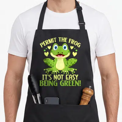 Permit frog aprons - Cartoon green frog with yellow hearts and stacked text reading PERMIT THE FROG IT'S NOT EASY BEING — shown on a black cotton canvas apron against a white background
