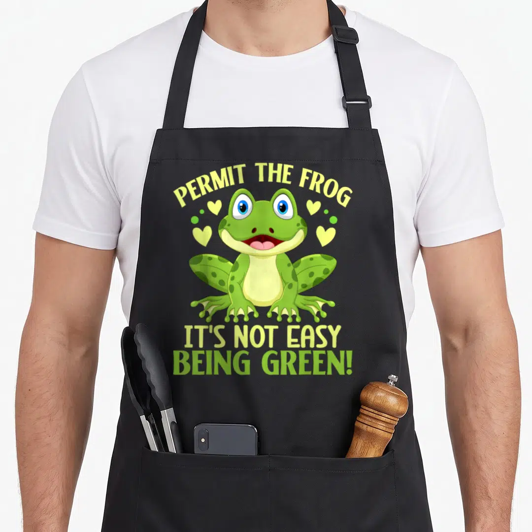 Permit frog aprons - Cartoon green frog with yellow hearts and stacked text reading PERMIT THE FROG IT'S NOT EASY BEING — shown on a black cotton canvas apron against a white background