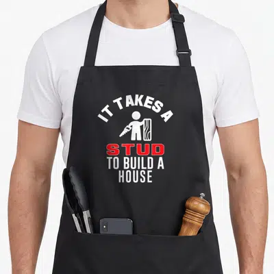 Carpenter builder building apron - White stick figure holding saw and wood next to red STUD text — shown on a black cotton canvas apron against a white background