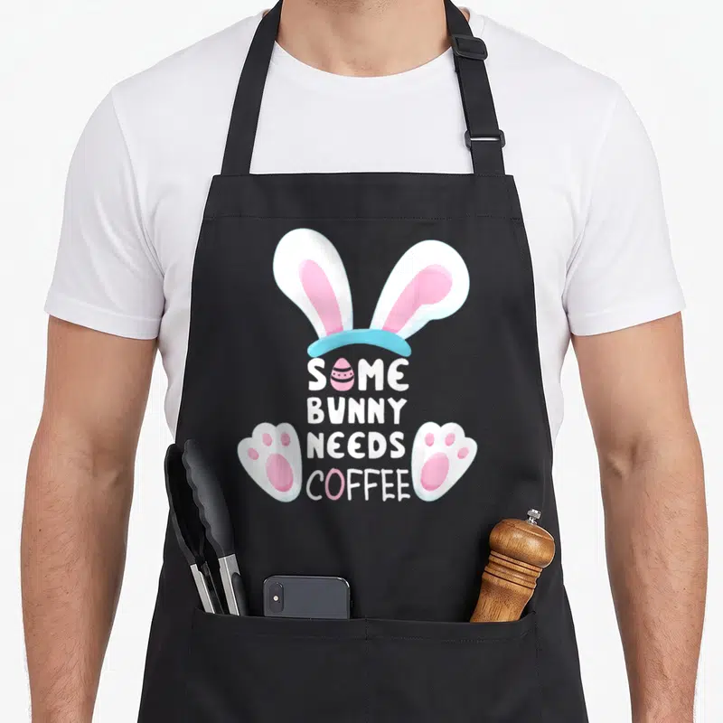 Coffee easter bunny aprons - Bunny ears and paws frame stacked text reading SOME BUNNY NEEDS COFFEE with a pink egg — shown on a black cotton canvas apron against a white background