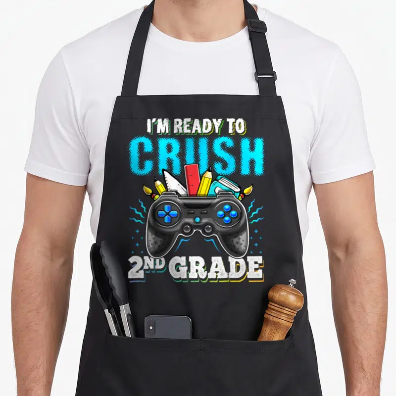 Crush video game apron - I'M READY TO CRUSH 2ND GRADE text over game controller with pencils and ruler — shown on a black cotton canvas apron against a white background