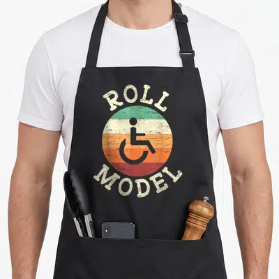 Role wheelchair apron - Retro ROLL MODEL text around striped circle with grey wheelchair icon — shown on a black cotton canvas apron against a white background