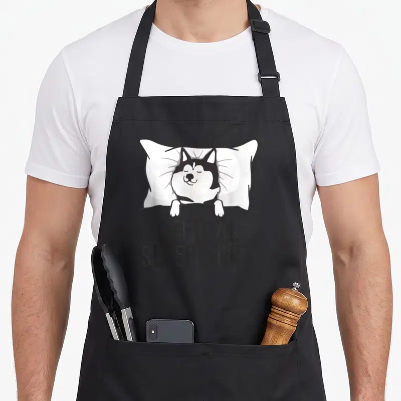 Pajama dog apron - Cartoon husky lying on white pillow under grey blanket with Offical Sleepshirt text — shown on a black cotton canvas apron against a white background