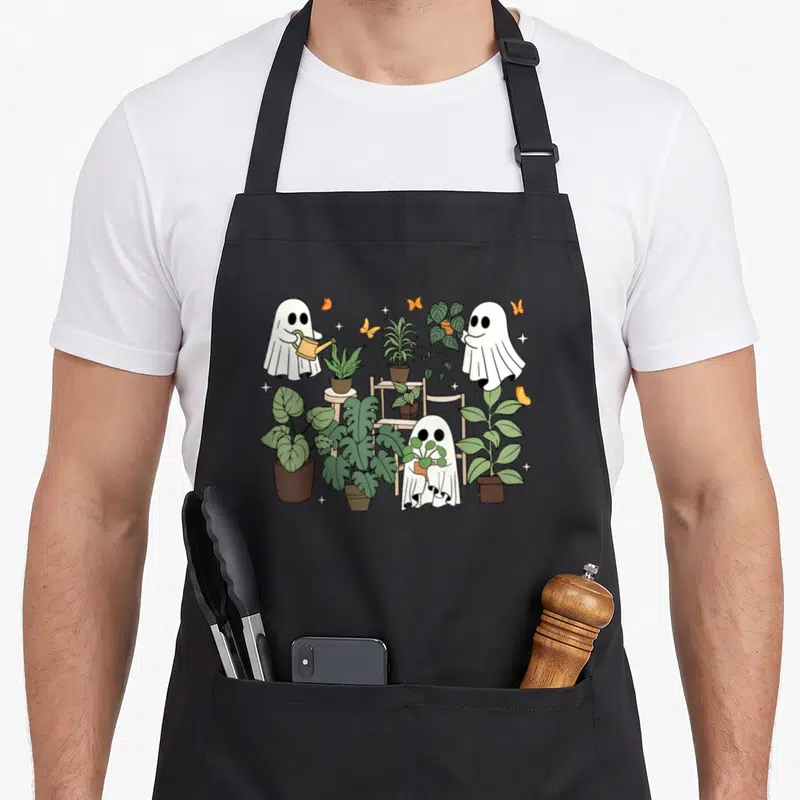 Ghost plant lady apron - Three cartoon ghosts holding plants and a watering can among green foliage and orange — shown on a black cotton canvas apron against a white background