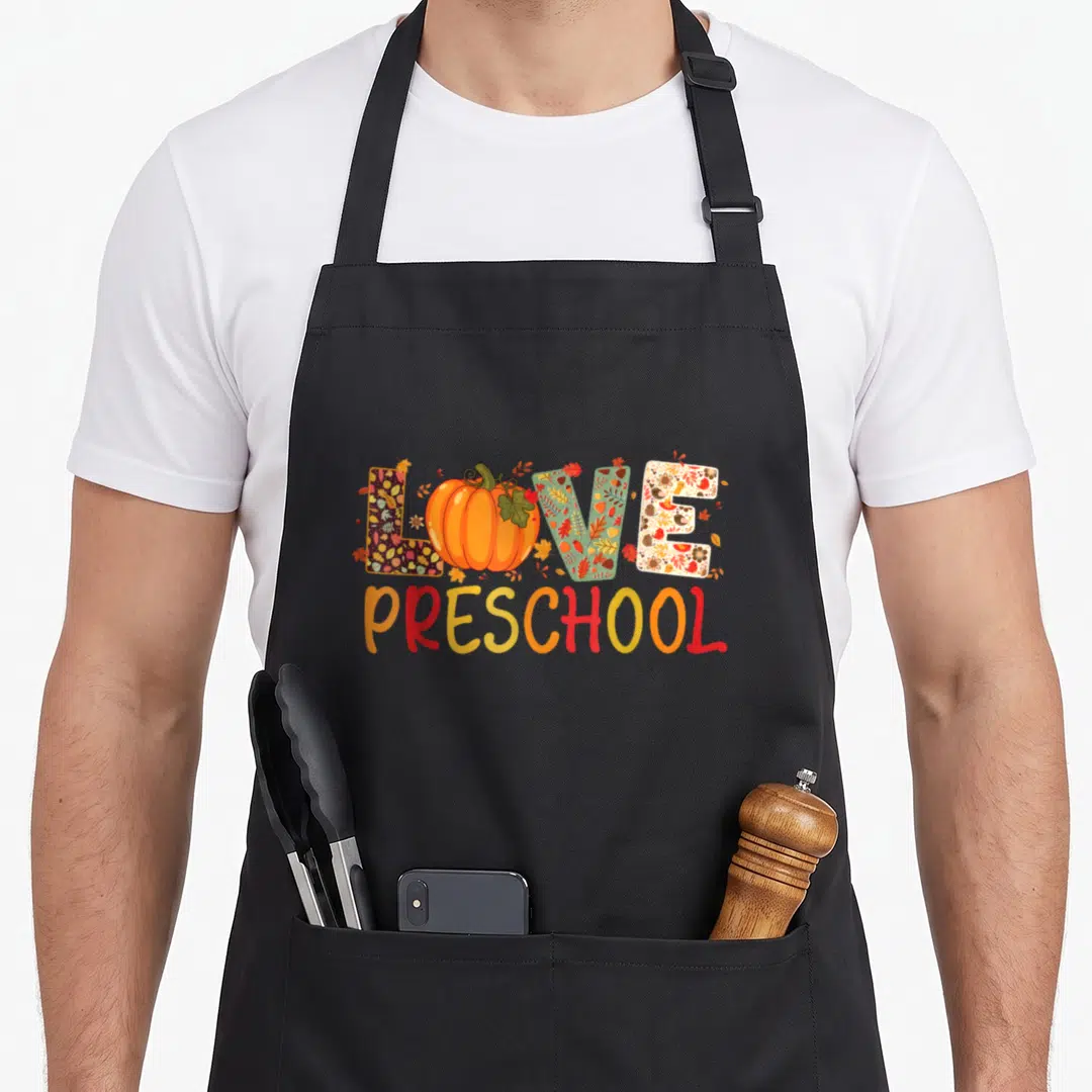 Fall preschool teacher apron - LOVE split by pumpkin with autumn-patterned letters, gradient PRESCHOOL below, cartoon — shown on a black cotton canvas apron against a white background