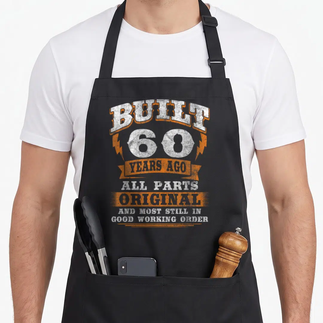 Built 60th birthday apron - Distressed white and orange text reading Built 60 Years Ago All Parts Original Good Working — shown on a black cotton canvas apron against a white background