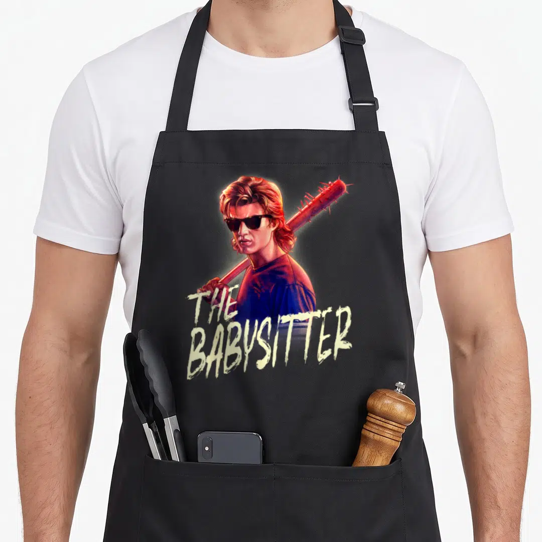 Steve stranger apron - Realistic man holding spiked bat with The Babysitter text in pale yellow — shown on a black cotton canvas apron against a white background
