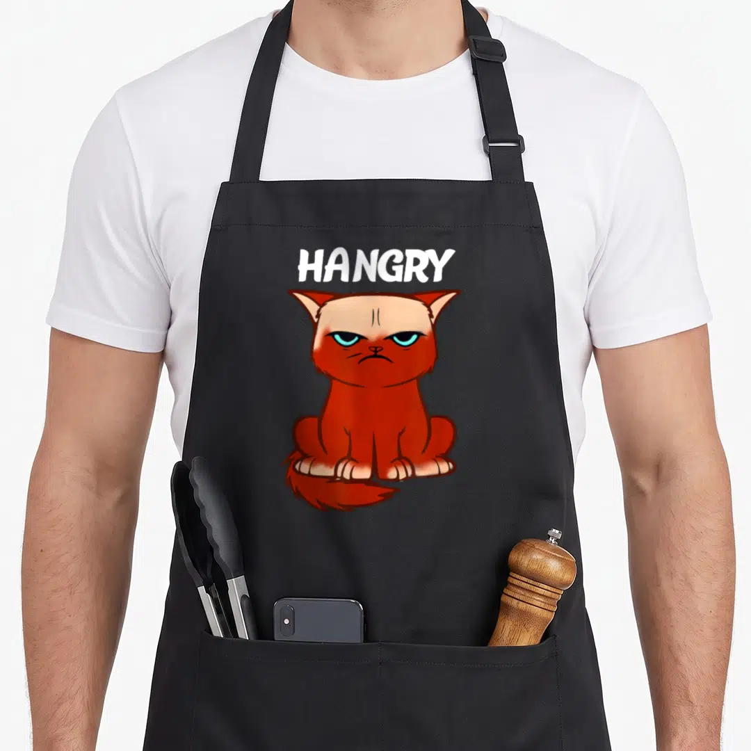 Hangry cat apron - White HANGRY text above a sitting angry orange cartoon cat illustration — shown on a black cotton canvas apron against a white background
