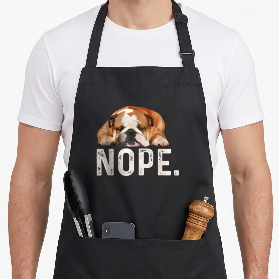 Nope lazy dog apron - Cartoon bulldog head resting on white NOPE text in brown and black — shown on a black cotton canvas apron against a white background