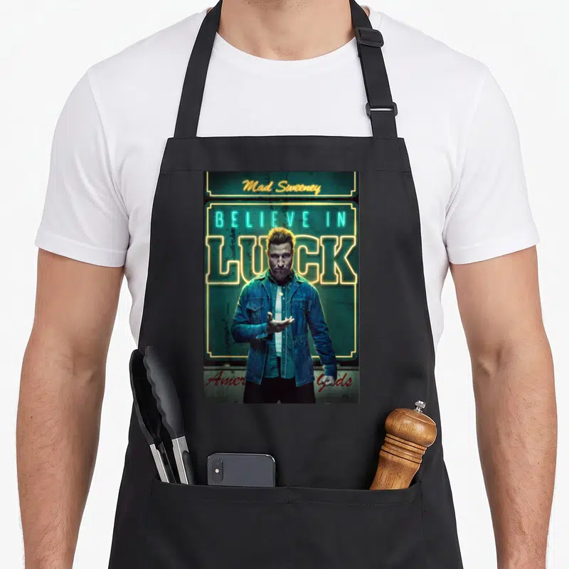 Mad english class apron - Man in denim jacket stands before neon sign reading BELIEVE IN LUCK and Mad Sweeney — shown on a black cotton canvas apron against a white background