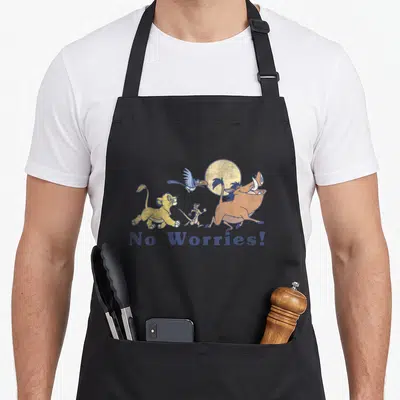 Worry lion dad apron - Cartoon lion cub, warthog, meerkat and bird walking in a row under a sun with blue No Worries — shown on a black cotton canvas apron against a white background