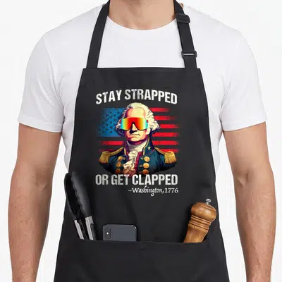 Clapped 4th of july apron - George Washington in rainbow sunglasses before distressed flag with Stay Strapped Or Get — shown on a black cotton canvas apron against a white background