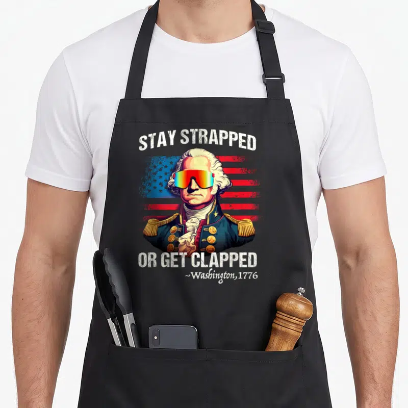 Clapped 4th of july apron - George Washington in rainbow sunglasses before distressed flag with Stay Strapped Or Get — shown on a black cotton canvas apron against a white background