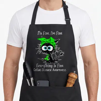 Celiac cat apron - Black scribbly monster with green ribbon and It's Fine text for Celiac awareness — shown on a black cotton canvas apron against a white background