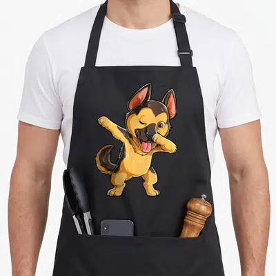 Dabbing dog mom apron - Cartoon tan and black German Shepherd standing on two legs dabbing with pink tongue out — shown on a black cotton canvas apron against a white background
