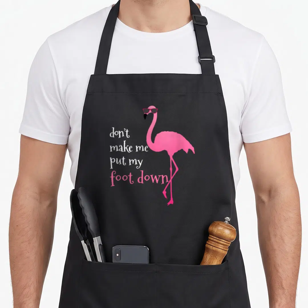 Cool foot flamingo apron - Pink flamingo with sunglasses and stacked text reading don't make me put my foot down — shown on a black cotton canvas apron against a white background