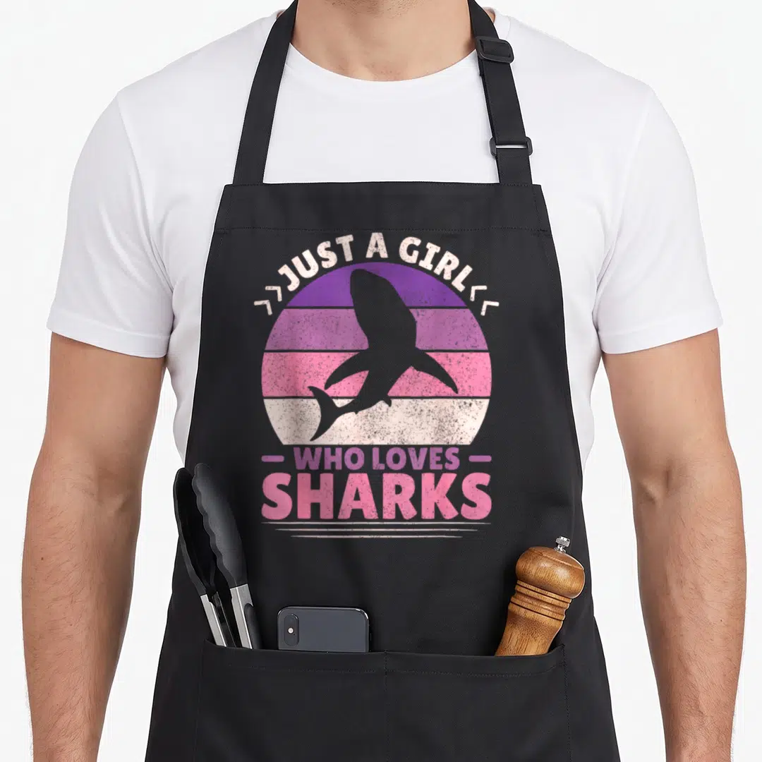 Vintage shark apron - Retro striped circle with grey shark silhouette and text Just A Girl Who Loves Sharks — shown on a black cotton canvas apron against a white background