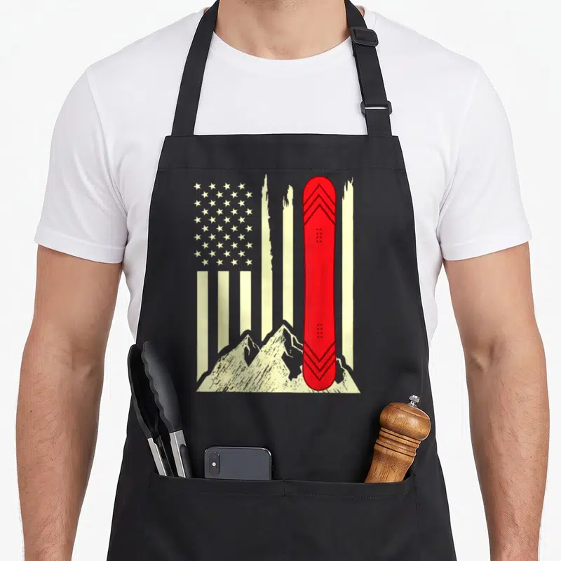 Snowboarding american flag apron - Red snowboard standing vertically before cream stars and stripes with mountain peaks — shown on a black cotton canvas apron against a white background