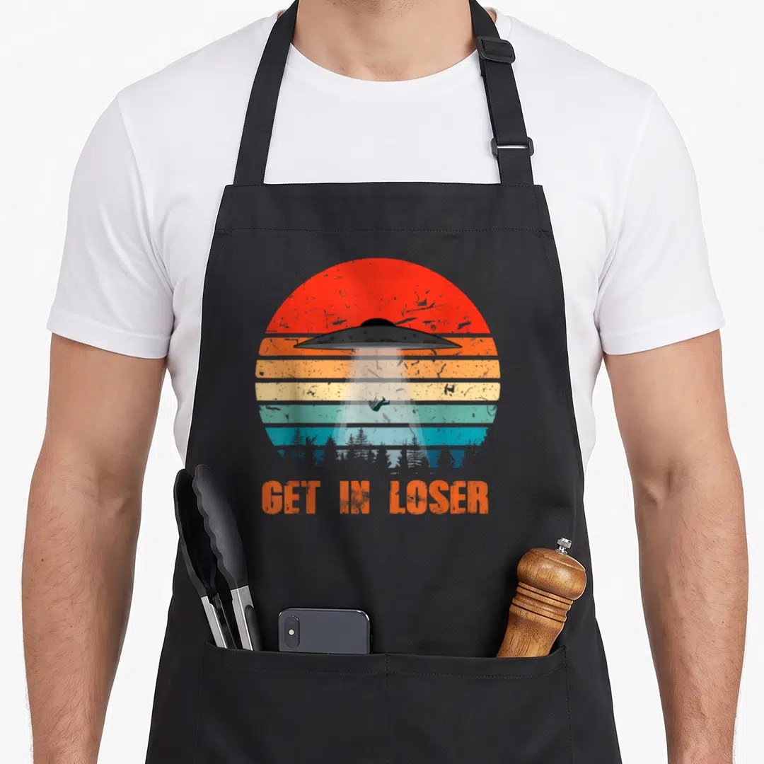 Loser conspiracy apron - Retro UFO beam over cow and trees with GET IN LOSER text on striped sun — shown on a black cotton canvas apron against a white background