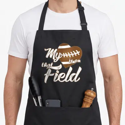 Funny field football apron - Brown football heart with white stripes and text My is on that Field — shown on a black cotton canvas apron against a white background
