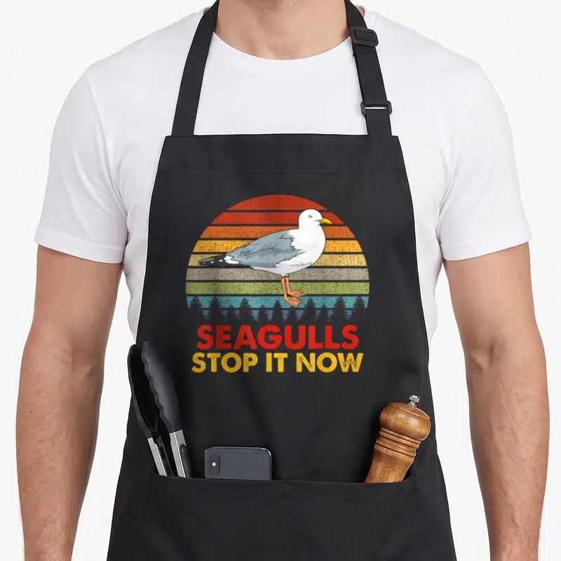 Seagull bird apron - Retro grey seagull standing before red yellow grey blue striped semi-circle with stacked text — shown on a black cotton canvas apron against a white background