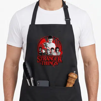 Growing stranger apron - Six character portraits in red circle above distressed STRANGER THINGS text — shown on a black cotton canvas apron against a white background