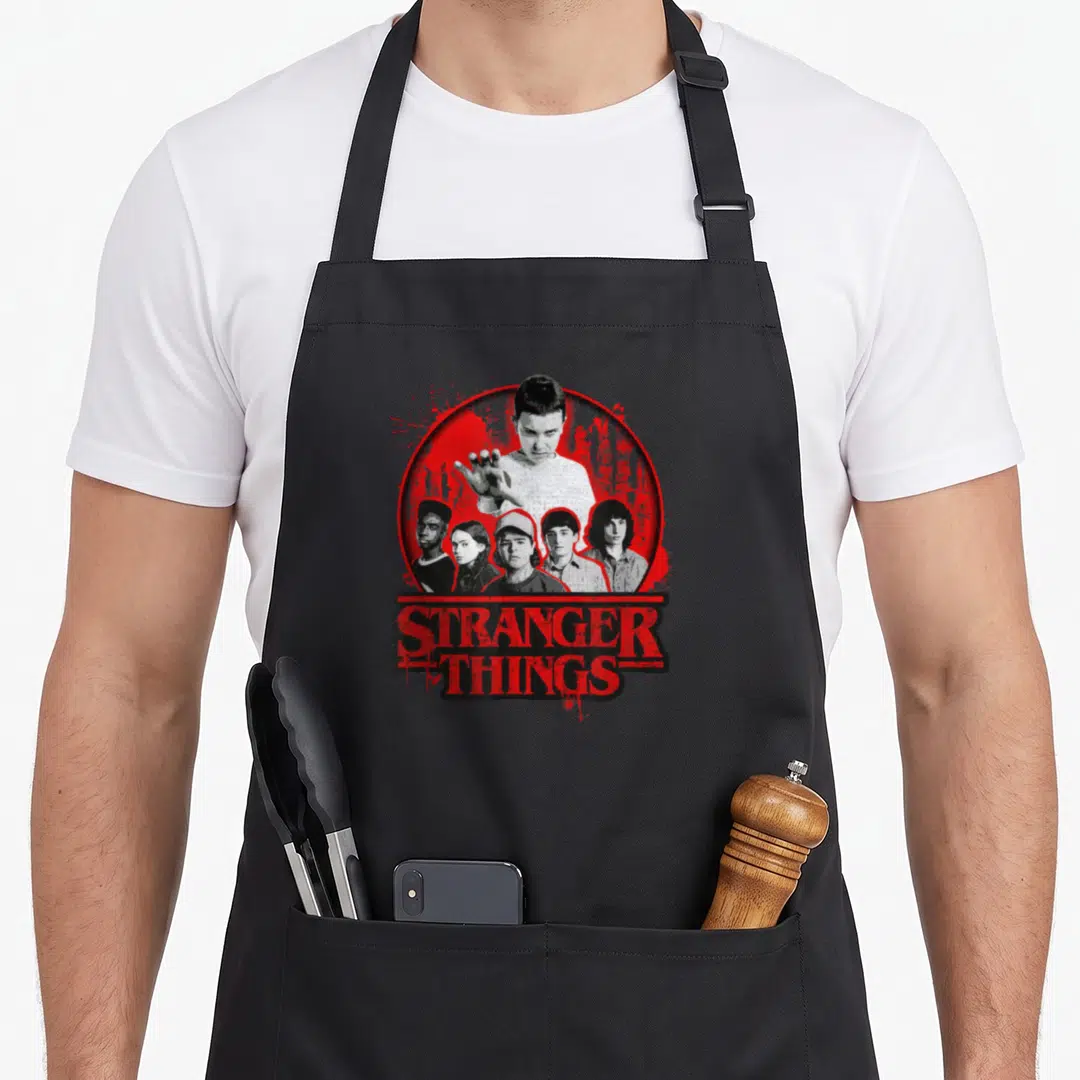 Growing stranger apron - Six character portraits in red circle above distressed STRANGER THINGS text — shown on a black cotton canvas apron against a white background