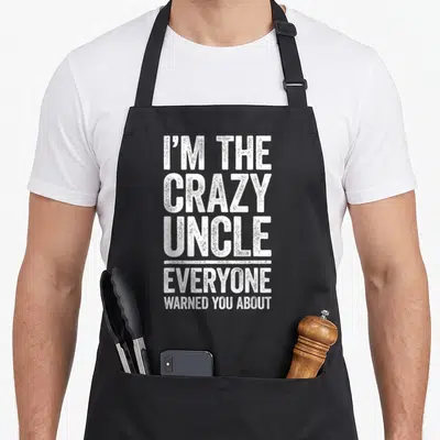 Uncle warned novelty apron - White stacked text I'M THE CRAZY UNCLE EVERYONE WARNED YOU ABOUT — shown on a black cotton canvas apron against a white background