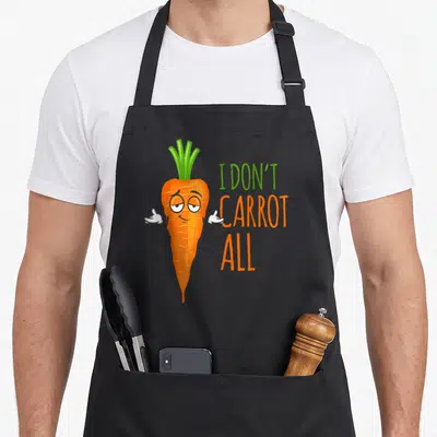Carrot vegan apron - Cartoon orange carrot shrugging next to stacked I Don't Carrot All text — shown on a black cotton canvas apron against a white background